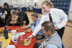 A student serves a plate of cheese to a table of seated adults.