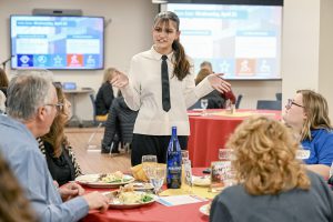A student in server attire converses with a table of seated adults.