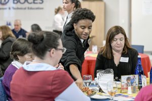 A student in an apron serves adults food at a table.