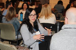 Two people at a conference are talking. One has there back to the camera, the other is gesturing with her hands