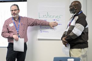 Two presenters are standing in front of a white board; one is gesturing to what's written. It says "listener." The other person is looking on
