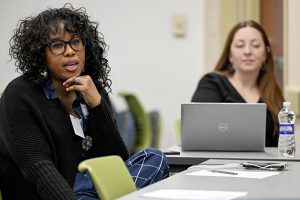 Two women are sitting at a table. One has her hand to her chin. She has curly hair