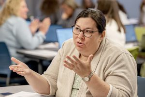 Conference attendee is gesturing with her hands. She is wearing a tan sweater, a wristwatch and glasses