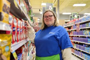A close-up photo of Skylar, a SELFI Program student.  She is wearing a blue Capital Region BOCES shirt and poses for a picture in front of a shelf of food as she completes her work-based learning hours at Price Chopper.