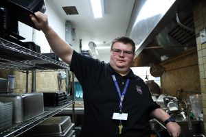 SELFI Program graduate Michael Sirigiano reaches for a container on top of rack as he cleans appliances during his shift at the Troy High School cafeteria.