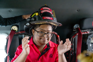 A Maywood Academy student wearing a red shirt takes a photo inside a fire truck while wear a Verdoy Firefighter helmet. 