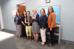 A group stands in front of a bulletin board. The recipient of the Extra Mile Award holds a plaque. She is flanked by five others.