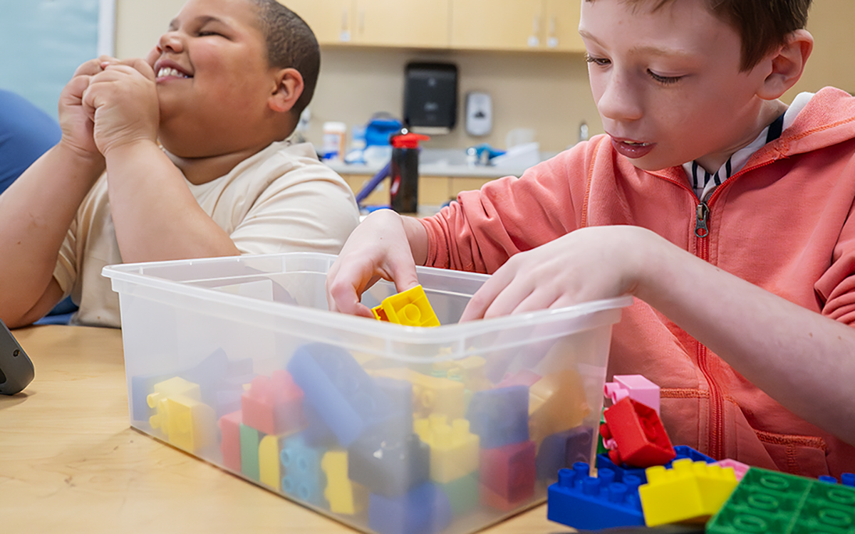 Students on the first day of school at Maywood Academy. Two students are seated together at a desk; one is looking up, smiling. The other is removing Legos from a clear plastic bin.