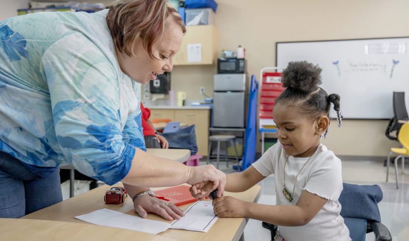 Two smiling teachers hand a student wearing a graduation cap and gown her diploma.
