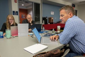 Three people, sitting at a table, are working on laptops. There are notebooks, water bottles, coffee cups and glasses on the table