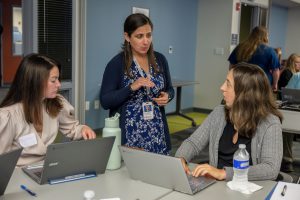 Two people are sitting at a table, working on laptops. Between them a woman is standing speaking and gesturing