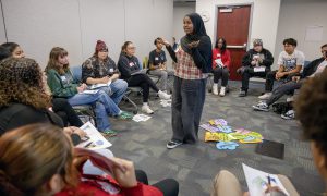 A student facilitator stands in front her peers as they're seated in a circle. She encourages them to use a set of prompts which are placed on the ground behind her.