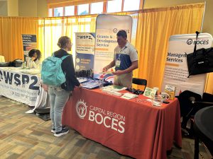 A person wearing a turquoise colored backpack speaks to a man standing behind a table with a red tablecloth that reads "Capital Region BOCES"