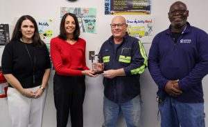 Four people are standing in a row, smiling at the camera. One is being presented with an award.