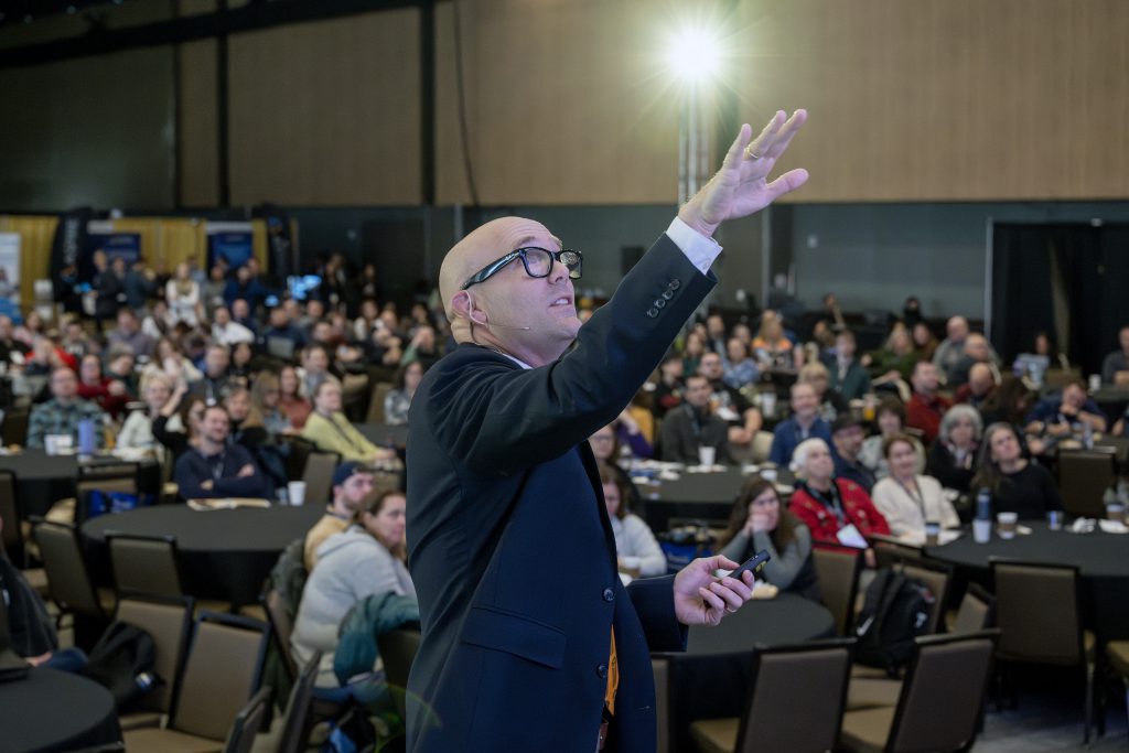 A man is speaking in front a crowd. He is gesturing towards the ceiling