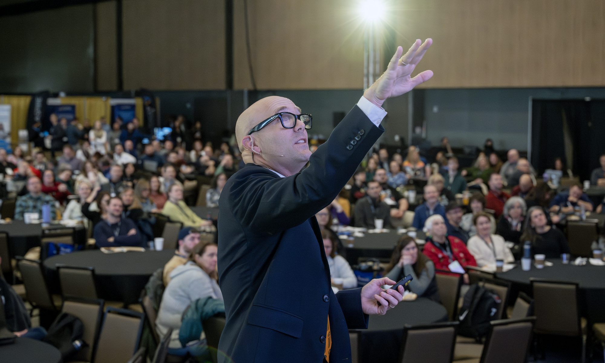 A man is speaking in front a crowd. He is gesturing towards the ceiling