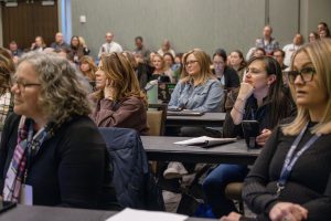 Attendees are seated at tables listening to a speaker