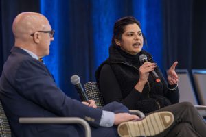 Two people are sitting on a stage, speaking into microphones. One is gesturing with her hand