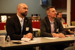 Two people are sitting side by at a table in a conference room listening to a speaker