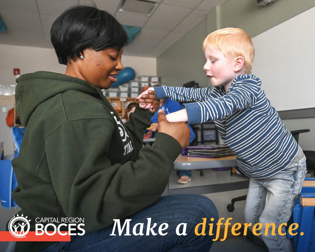A teaching assistant is photographed while helping a young student stand from his seat.