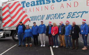 SELFI Program students and staff wearing their blue CRB BOCES shirts and Santa hats, pose for a photo in front of a large white Teamsters truck with representatives from Teamsters and NYSUT.