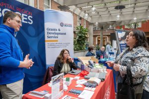 Capital Region BOCES staff engage in conversation with event attendees at a Grants & Development Service booth, surrounded by brochures, promotional items, and informational signage.