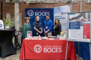 Four BOCES staff stand behind a table with a red table cloth that reads Capital Region BOCES. Behind the people are backdrops with the BOCES logo and type that reads Capital Region BOCES and Grants & Development Service. Next to the table is a poster on an easel with the headline Municipal Services Information Session