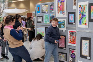A student smiles while pointing to his artwork as his photo is taken by a family member.