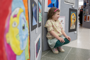 A photo of a young student as she kneels down next her artwork that's on display.  