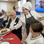 A student wearing white culinary uniform and a blue apron serves a plate of food to a man seated at a table