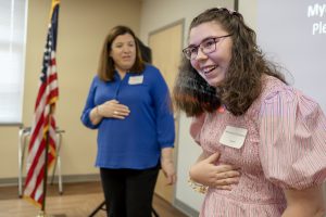 A student wearing a pink dress smiles, standing next to a teacher waring a blue shirt