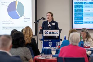 A woman is speaking at a podium into a microphone in front of people seated at tables. Behind her are graphs and charts projected onto screens