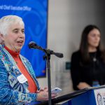 A woman wearing a blue sweater over an orange turtleneck speaks into a microphone at a podium