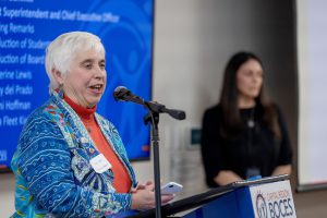 A woman wearing a blue sweater over an orange turtleneck speaks into a microphone at a podium