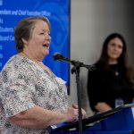 A woman wearing a white and black top speaks at a podium.