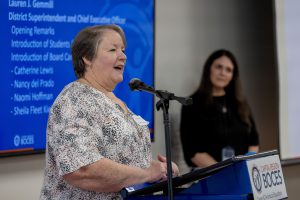 A woman wearing a white and black top speaks at a podium.
