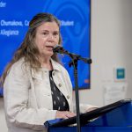 A woman speaks into a microphone. She is wearing a tan jacket and a dark shirt.