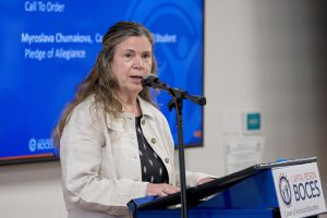 A woman speaks into a microphone. She is wearing a tan jacket and a dark shirt.