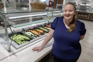A woman wearing a navy blue top is standing in a cafeteria, next to a salad bar. She is smiling at the camera