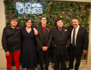 A group photo of three male students, Special Education Deputy Director Kristen Roys and Maywood Academy Principal Gabriele Barbato as they pose in front of a flower wall and a sign made of lights that reads "Duo at the Dunes."