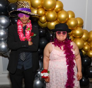 A photo of a male and female students as they pose for a photo in front of a sliver, black and gold balloons garland while wearing their festive costume props.