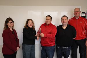 Five people are standing in a line in the center is a man wearing a red shirt, he is receiving an award from a woman on his left.