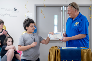 A young male student wearing a gray shirt accept his moving-up certification from his teacher, Mr. Darpino.