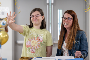 A female student wearing a yellow shirt stands beside her teacher at the podium. The student is waving to the audience as the teacher smiles.