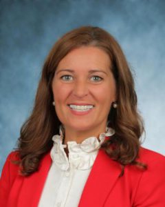 This is a professional head shot of a woman who is smiling at the camera. She is wearing a high neck white blouse and an orange blazer.
