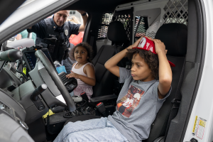 Two excited students sit in the front seats of a police car as the school resource officer looks in through the car window.