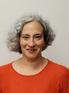In a headshot, a woman with grey wavy hair that is about chin length, wearing an orange top.