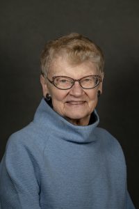 A woman is smiling at the camera wearing a slate blue high neck shirt. She is wearing glasses