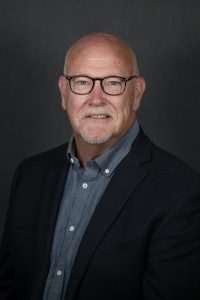 In this head shot, a man is wearing a dark suit coat and a medium blue button down shirt. He is looking at the camera and is wearing glasses