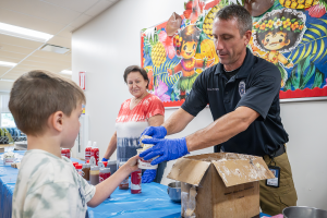 School Resource Officer Matt Stott hands a cup of ice cream to a young student as a teaching assistant smiles in the background.